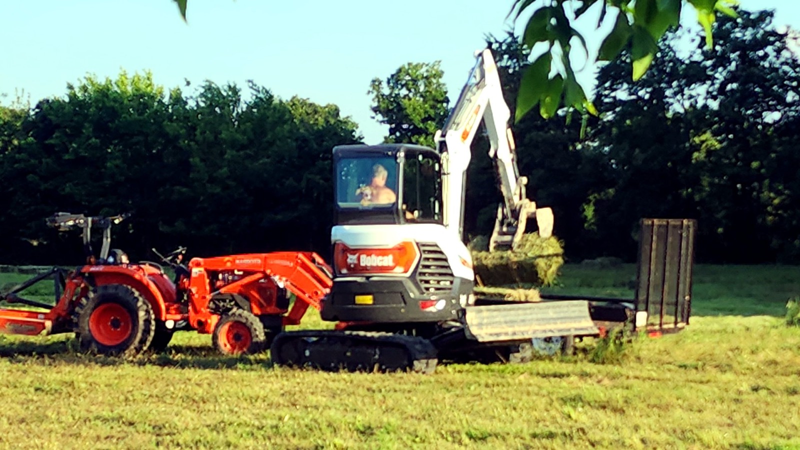 Compact excavator loading hay beside a tractor in a field